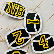 Load image into Gallery viewer, Hockey puck first birthday photo banner featuring 13 hockey pucks. Each hockey puck is sized to fit one 4x6 portrait or orientation photo. The numbers are yellow and the black. The number colors are customizable and can be matched to coordinate with your favorite hockey team colors.