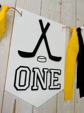 Load image into Gallery viewer, Hockey first birthday high chair banner featuring crossed hockey sticks, hockey puck, layered varsity font letters spelling the word "ONE" and yellow, black and white fabric strips. This hockey first birthday high chair banner is customizable, handcrafted and designed by Laura, the owner of Declan & Smith Party Decor.