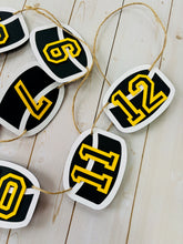 Load image into Gallery viewer, Hockey puck first birthday photo banner featuring 13 hockey pucks. Each hockey puck is sized to fit one 4x6 portrait or orientation photo. The numbers are yellow and the black. The number colors are customizable and can be matched to coordinate with your favorite hockey team colors.