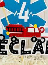 Load image into Gallery viewer, The first responders cake topper features a police car, firetruck, ambulance, road letters, EMT emblem with age and heart beat monitor with a yellow siren.