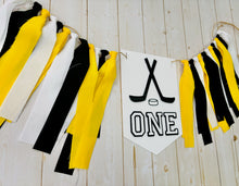 Load image into Gallery viewer, Hockey first birthday high chair banner featuring crossed hockey sticks, hockey puck, layered varsity font letters spelling the word "ONE" and yellow, black and white fabric strips. This hockey first birthday high chair banner is customizable, handcrafted and designed by Laura, the owner of Declan & Smith Party Decor.