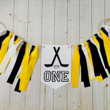 Load image into Gallery viewer, Hockey first birthday high chair banner featuring crossed hockey sticks, hockey puck, layered varsity font letters spelling the word "ONE" and yellow, black and white fabric strips. This hockey first birthday high chair banner is customizable, handcrafted and designed by Laura, the owner of Declan & Smith Party Decor.