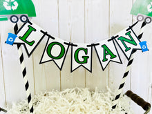 Load image into Gallery viewer, Recycling trash truck cake topper featuring two green trash trucks, pennant with green letters strung between black and white striped paper straws. There are two blue recycling trash bins on the beginning and end letters on the cake bunting cake topper.
