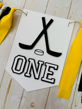 Load image into Gallery viewer, Hockey first birthday high chair banner featuring crossed hockey sticks, hockey puck, layered varsity font letters spelling the word "ONE" and yellow, black and white fabric strips. This hockey first birthday high chair banner is customizable, handcrafted and designed by Laura, the owner of Declan & Smith Party Decor.