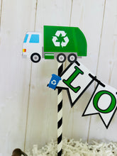 Load image into Gallery viewer, Recycling trash truck cake topper featuring two green trash trucks, pennant with green letters strung between black and white striped paper straws. There are two blue recycling trash bins on the beginning and end letters on the cake bunting cake topper.
