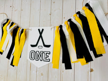 Load image into Gallery viewer, Hockey first birthday high chair banner featuring crossed hockey sticks, hockey puck, layered varsity font letters spelling the word "ONE" and yellow, black and white fabric strips. This hockey first birthday high chair banner is customizable, handcrafted and designed by Laura, the owner of Declan & Smith Party Decor.