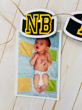 Load image into Gallery viewer, Hockey puck first birthday photo banner featuring 13 hockey pucks. Each hockey puck is sized to fit one 4x6 portrait or orientation photo. The numbers are yellow and the black. The number colors are customizable and can be matched to coordinate with your favorite hockey team colors.