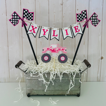 Load image into Gallery viewer, Pink monster truck cake topper. The cake topper is customizable and can display an age, name, word or phrase. The colors are also customizable. The pink monster truck cake topper features two checkered flags, pennant banner and a monster truck to display underneath the bunting. The pink monster truck cake topper is designed and handcrafted by Laura, the owner of Declan & Smith Party Decor.