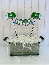 Load image into Gallery viewer, Recycling trash truck cake topper featuring two green trash trucks, pennant with green letters strung between black and white striped paper straws. There are two blue recycling trash bins on the beginning and end letters on the cake bunting cake topper.