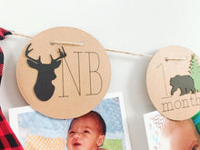 Load image into Gallery viewer, Lumberjack first birthday banner featuring deer head, bear, wolves and evergreen trees. The photo banner includes 13 circles to display your child's first year milestone photos. There are two strips of red and black buffalo plaid fabric on each end of the banner.
