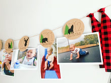 Load image into Gallery viewer, Lumberjack first birthday banner featuring deer head, bear, wolves and evergreen trees. The photo banner includes 13 circles to display your child's first year milestone photos. There are two strips of red and black buffalo plaid fabric on each end of the banner.