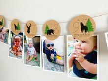 Load image into Gallery viewer, Lumberjack first birthday banner featuring deer head, bear, wolves and evergreen trees. The photo banner includes 13 circles to display your child's first year milestone photos. There are two strips of red and black buffalo plaid fabric on each end of the banner.