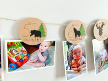 Load image into Gallery viewer, Lumberjack first birthday banner featuring deer head, bear, wolves and evergreen trees. The photo banner includes 13 circles to display your child's first year milestone photos. There are two strips of red and black buffalo plaid fabric on each end of the banner.