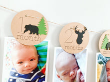 Load image into Gallery viewer, Lumberjack first birthday banner featuring deer head, bear, wolves and evergreen trees. The photo banner includes 13 circles to display your child's first year milestone photos. There are two strips of red and black buffalo plaid fabric on each end of the banner.