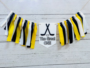 Hockey first birthday high chair banner featuring crossed hockey sticks, hockey puck, layered varsity font letters spelling the word "ONE" and yellow, black and white fabric strips. This hockey first birthday high chair banner is customizable, handcrafted and designed by Laura, the owner of Declan & Smith Party Decor.