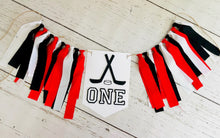 Load image into Gallery viewer, Hockey first birthday high chair banner featuring crossed hockey sticks, hockey puck, layered varsity font letters spelling the word &quot;ONE&quot; and red, black and white fabric strips. This hockey first birthday high chair banner is customizable, handcrafted and designed by Laura, the owner of Declan &amp; Smith Party Decor.
