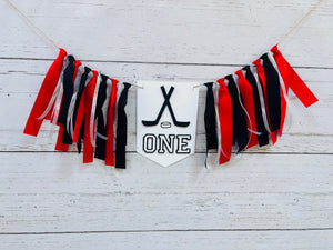 Hockey first birthday high chair banner featuring crossed hockey sticks, hockey puck, layered varsity font letters spelling the word "ONE" and red, black and silver fabric strips. This hockey first birthday high chair banner is customizable, handcrafted and designed by Laura, the owner of Declan & Smith Party Decor.
