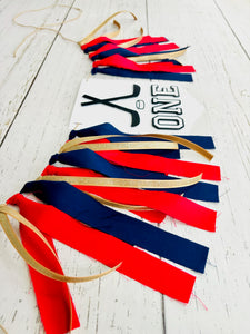 Hockey first birthday high chair banner featuring crossed hockey sticks, hockey puck, layered varsity font letters spelling the word "ONE" and red, navy blue and gold fabric strips. This hockey first birthday high chair banner is customizable, handcrafted and designed by Laura, the owner of Declan & Smith Party Decor.