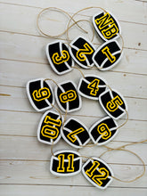 Load image into Gallery viewer, Hockey puck first birthday photo banner featuring 13 hockey pucks. Each hockey puck is sized to fit one 4x6 portrait or orientation photo. The numbers are yellow and the black. The number colors are customizable and can be matched to coordinate with your favorite hockey team colors. 
