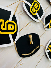 Load image into Gallery viewer, Hockey puck first birthday photo banner featuring 13 hockey pucks. Each hockey puck is sized to fit one 4x6 portrait or orientation photo. The numbers are yellow and the black. The number colors are customizable and can be matched to coordinate with your favorite hockey team colors. 
