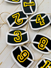 Load image into Gallery viewer, Hockey puck first birthday photo banner featuring 13 hockey pucks. Each hockey puck is sized to fit one 4x6 portrait or orientation photo. The numbers are yellow and the black. The number colors are customizable and can be matched to coordinate with your favorite hockey team colors. 

