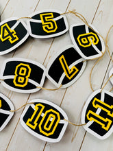 Load image into Gallery viewer, Hockey puck first birthday photo banner featuring 13 hockey pucks. Each hockey puck is sized to fit one 4x6 portrait or orientation photo. The numbers are yellow and the black. The number colors are customizable and can be matched to coordinate with your favorite hockey team colors. 
