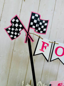 Pink monster truck cake topper. The cake topper is customizable and can display an age, name, word or phrase. The colors are also customizable. The pink monster truck cake topper features two checkered flags, pennant banner and a monster truck to display underneath the bunting. The pink monster truck cake topper is designed and handcrafted by Laura, the owner of Declan & Smith Party Decor. 
