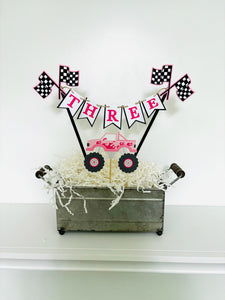 Pink monster truck cake topper. The cake topper is customizable and can display an age, name, word or phrase. The colors are also customizable. The pink monster truck cake topper features two checkered flags, pennant banner and a monster truck to display underneath the bunting. The pink monster truck cake topper is designed and handcrafted by Laura, the owner of Declan & Smith Party Decor. 