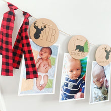 Load image into Gallery viewer, Lumberjack first birthday banner featuring deer head, bear, wolves and evergreen trees. The photo banner includes 13 circles to display your child&#39;s first year milestone photos. There are two strips of red and black buffalo plaid fabric on each end of the banner. 
